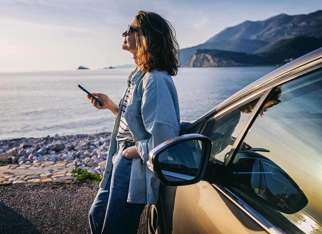 Service Center - Middle Aged Woman Wearing Sunglasses Standing Next to her Car While Holding her Phone on the Beach at Sunset